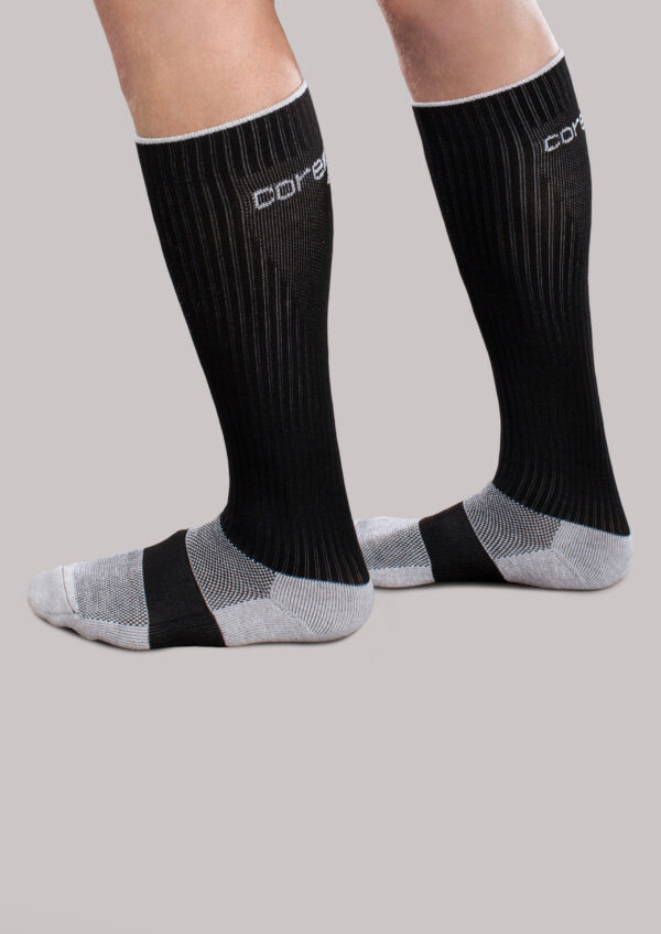 A person wearing Core-Sport by Therafirm knee-high black and gray compression socks, standing on a gray background.