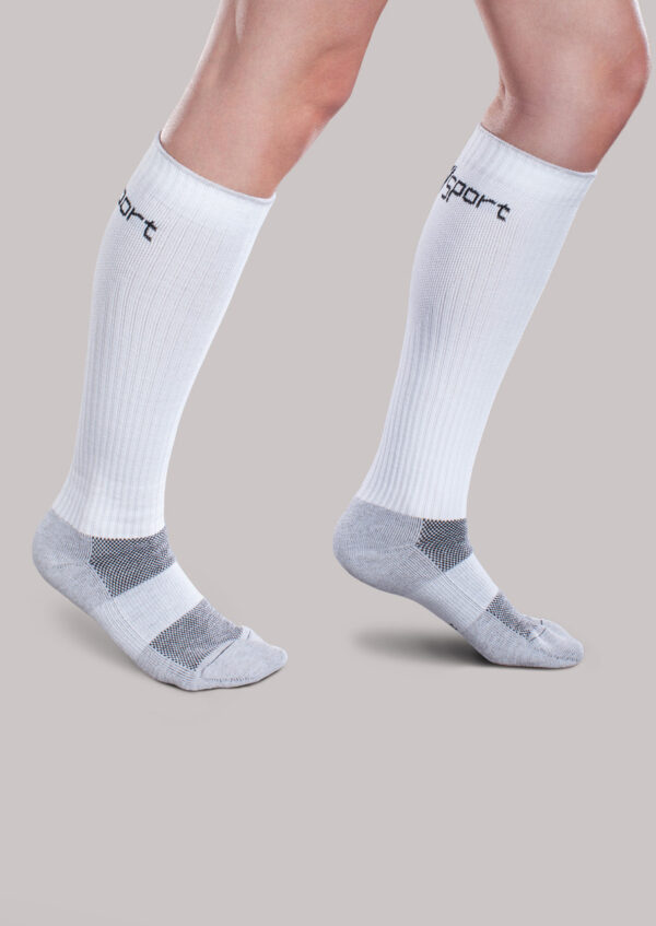 Two legs wearing Core-Sport by Therafirm white knee-high athletic socks with grey accents, standing on one leg against a plain grey background.